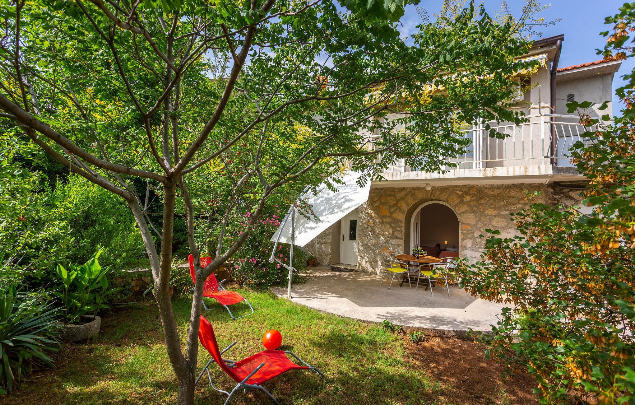 Un bâtiment en pierre doté d’un balcon, une terrasse avec une table et des chaises, et un jardin avec des arbres et des plantes.