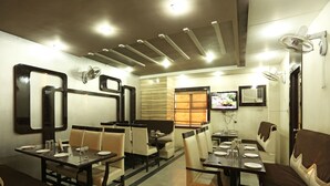 A restaurant with tables and chairs, a TV mounted on the wall, and ceiling fans. - OYO 1671 Hotel Sundaram (Prayagraj)
