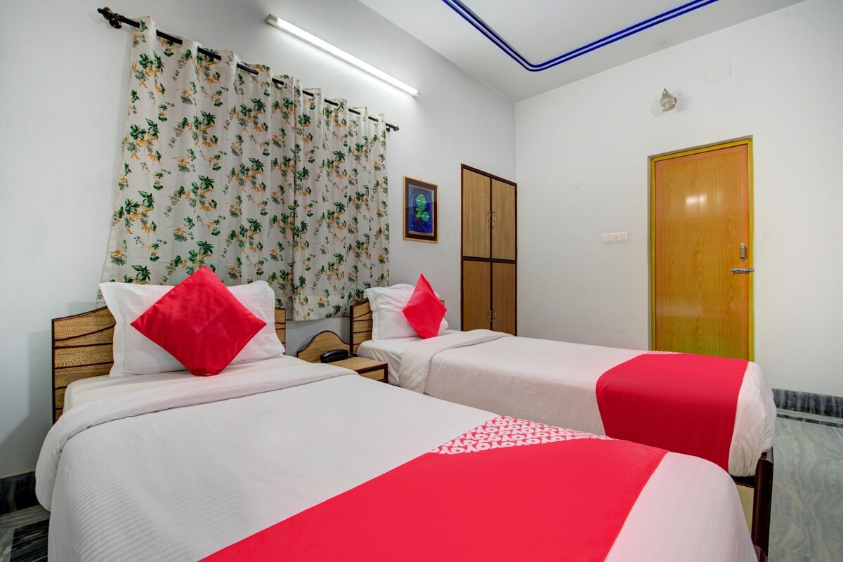 A hotel room with two beds, each with white bedding and red accents, a wooden wardrobe, a wooden door, and a window with floral curtains.