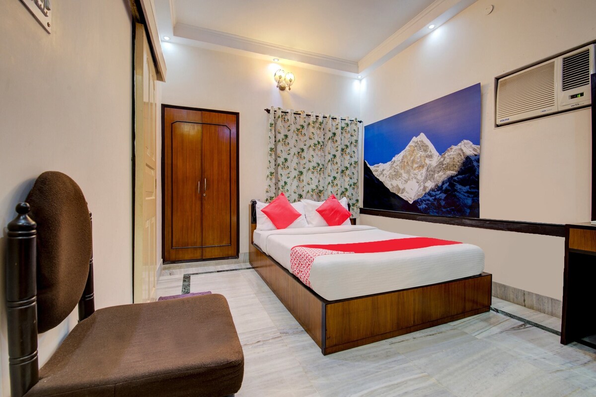 A hotel room with a bed, a wooden wardrobe, a bench, a wall-mounted air conditioner, and a painting of a mountain.