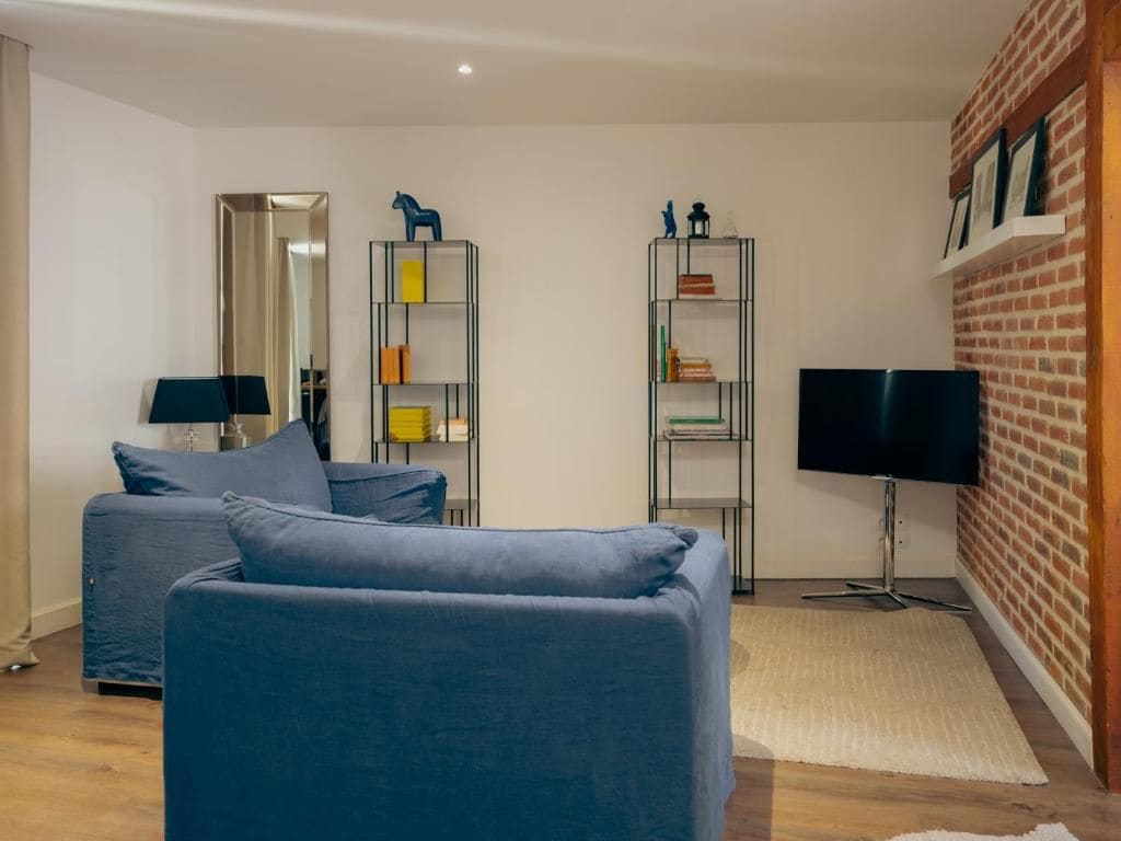 A modern living room with a sofa, a TV, and two shelving units against a brick wall.