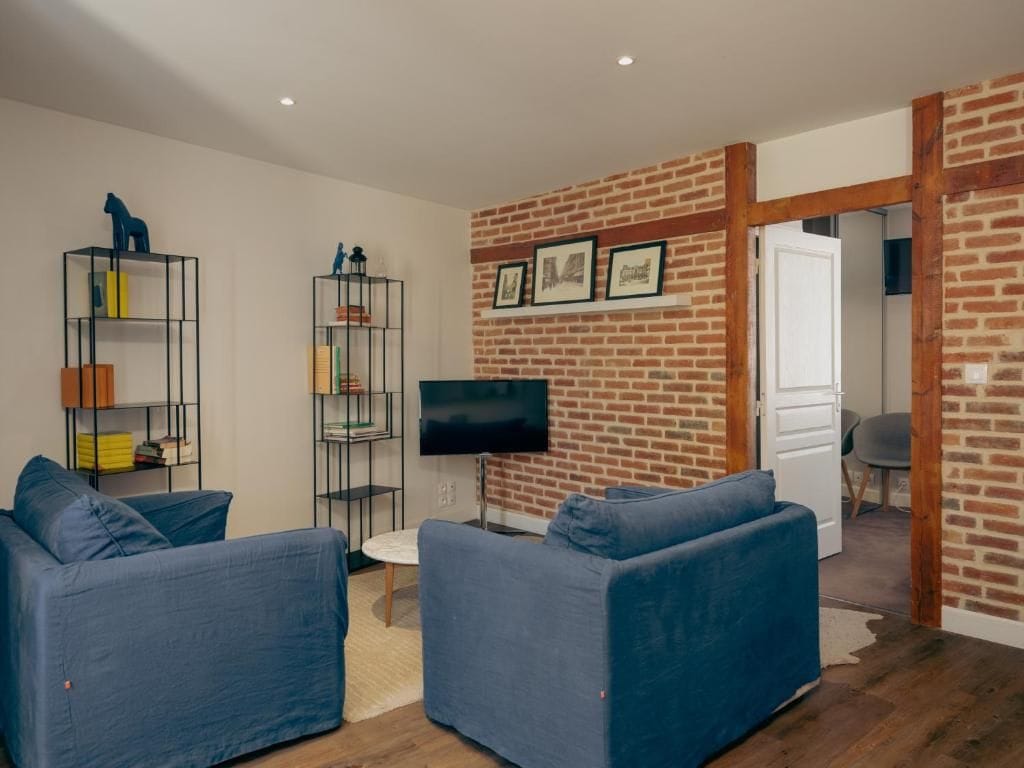 A living room with a brick wall, two blue sofas, a TV, and a bookshelf.