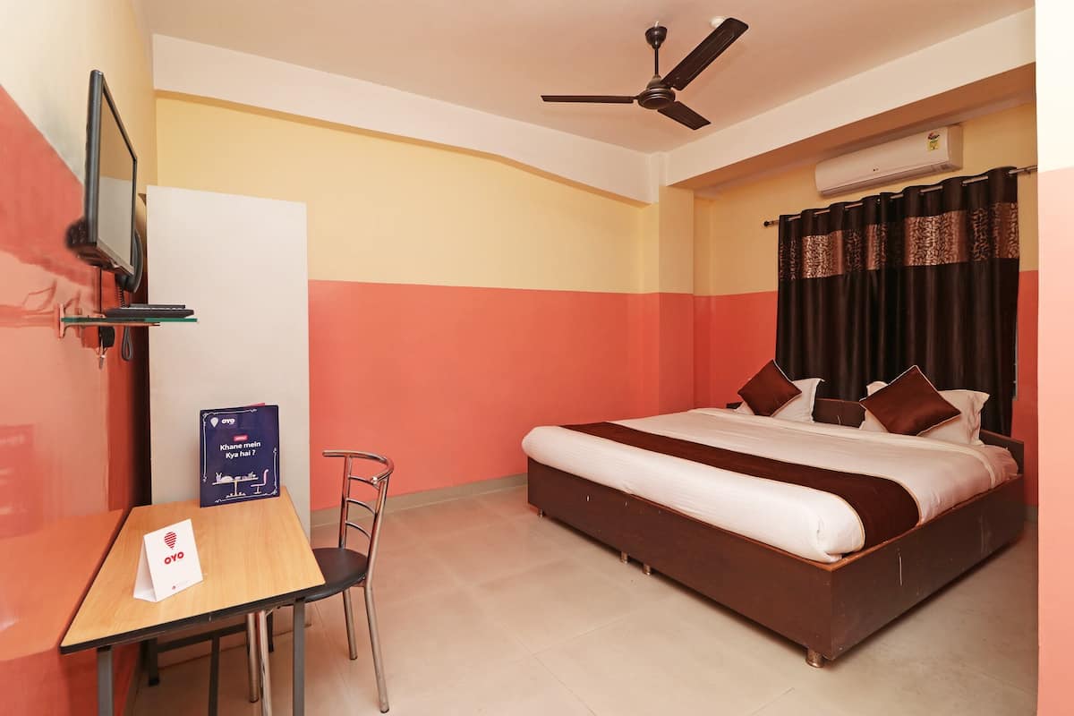 A hotel room with a bed, a desk, a chair, a TV mounted on the wall, and an air conditioning unit.