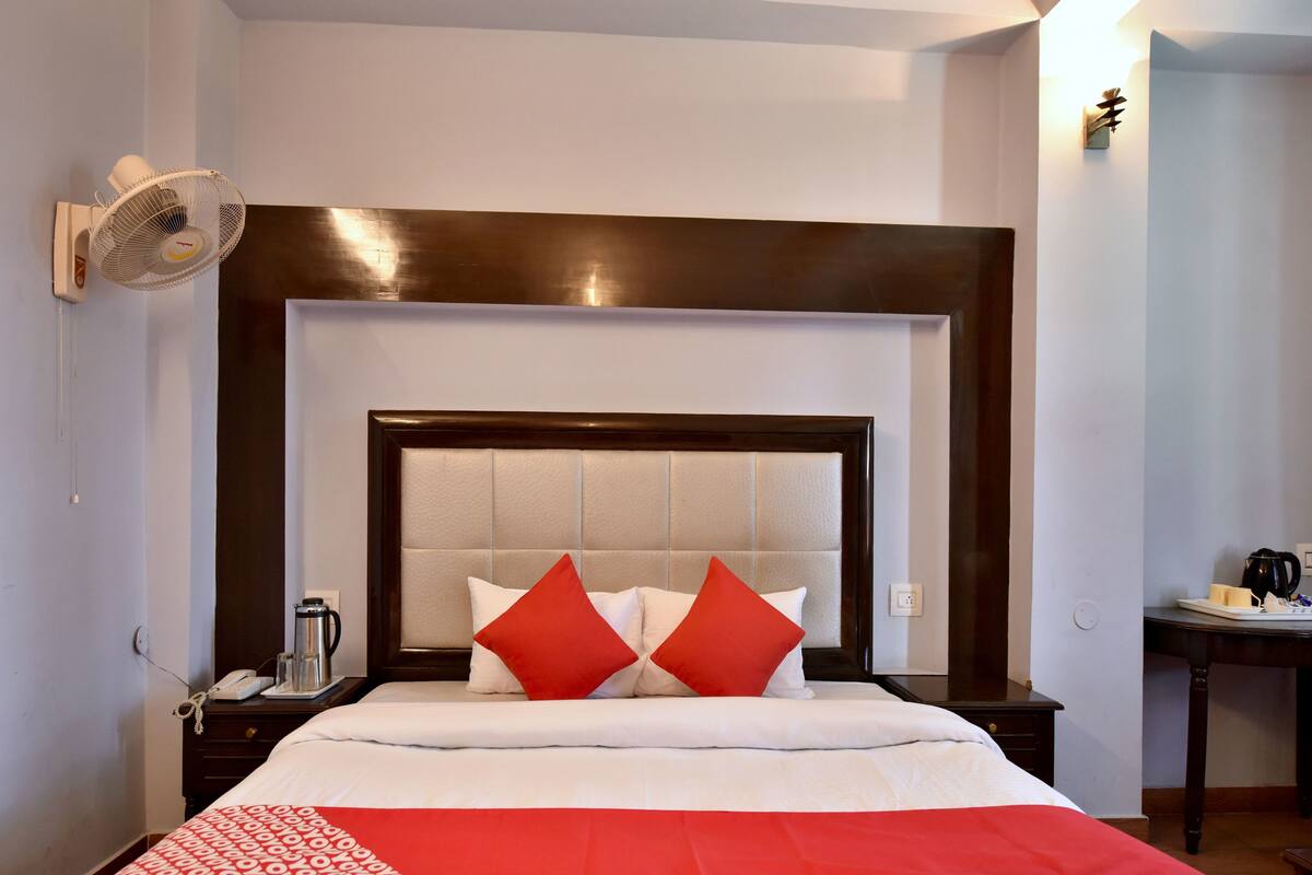 Una habitación de hotel con una cama grande, una mesita de noche con lámpara, un ventilador en la pared y una pequeña mesa con hervidor y tazas.