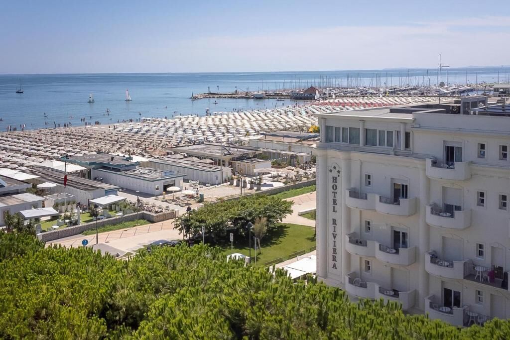 Ein Hotel an der Küste mit mehreren Balkonen, einer Marina mit Booten und einem Strandbereich mit Sonnenschirmen.