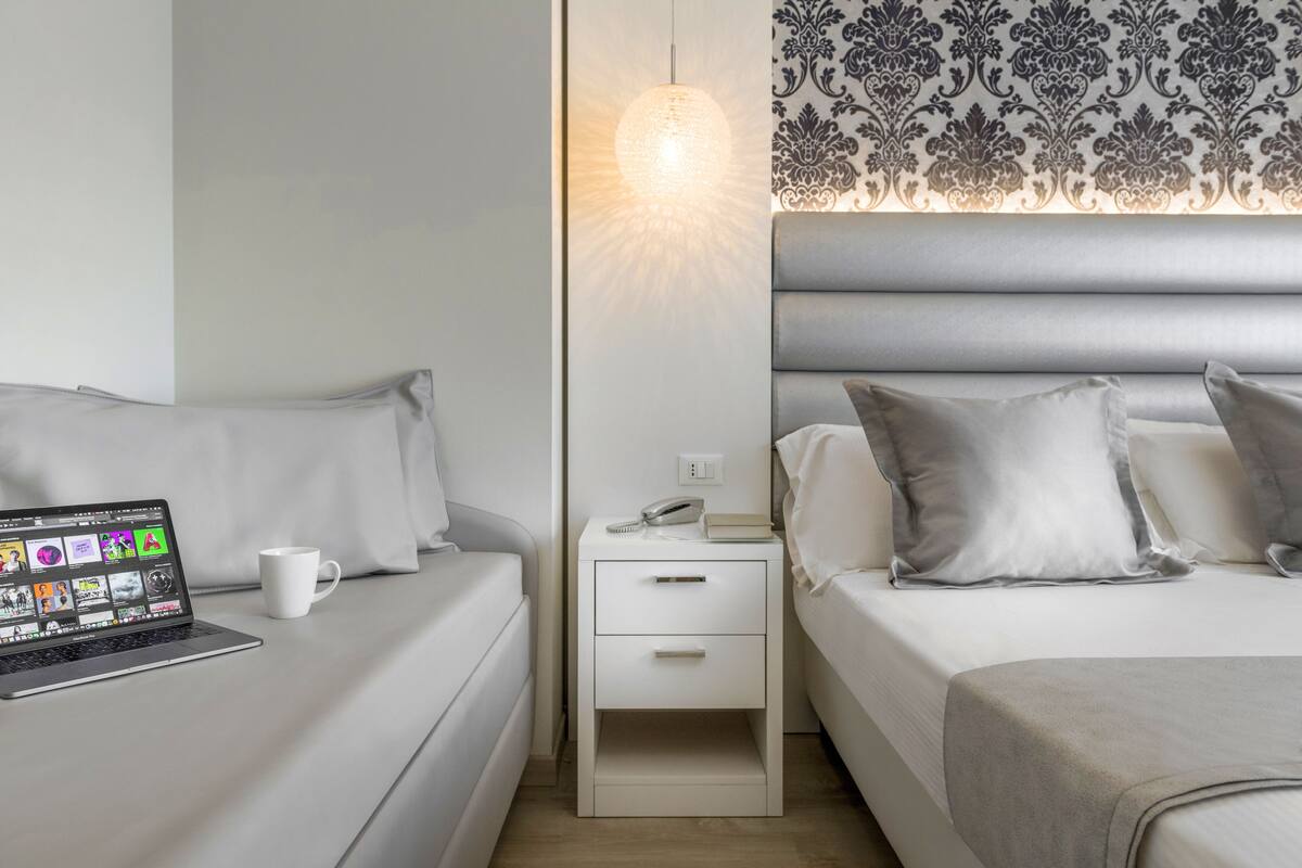 A modern hotel room with a bed, a bedside table with a lamp, a laptop, and a white mug.