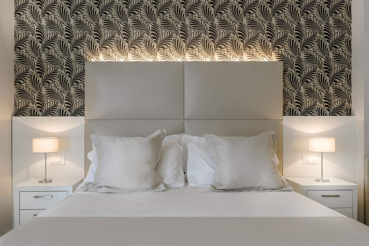 A neatly made bed with white linens and pillows, flanked by two bedside tables with lamps, set against a wall with a leaf-patterned wallpaper.