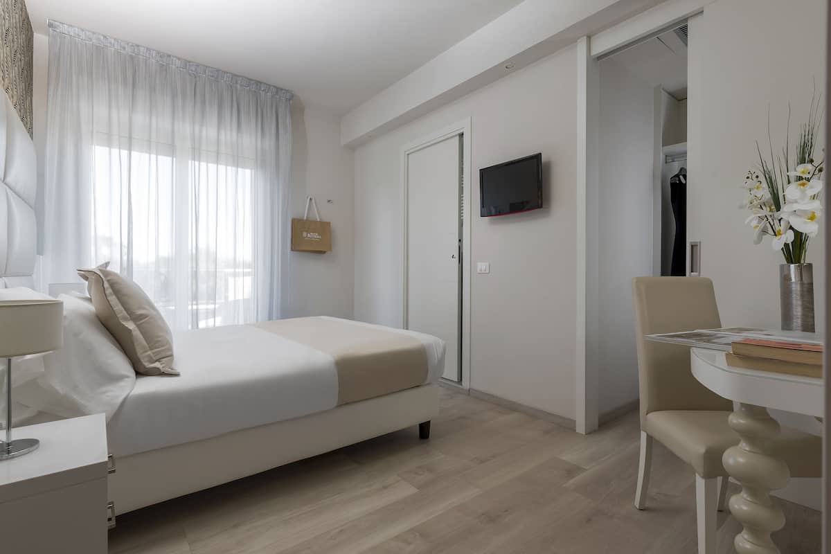 A modern hotel room with a bed, a TV mounted on the wall, a desk with a lamp, and a chair.