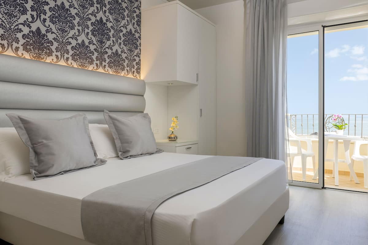 A modern hotel room with a large bed, a nightstand, a decorative wall, and a balcony with outdoor furniture.