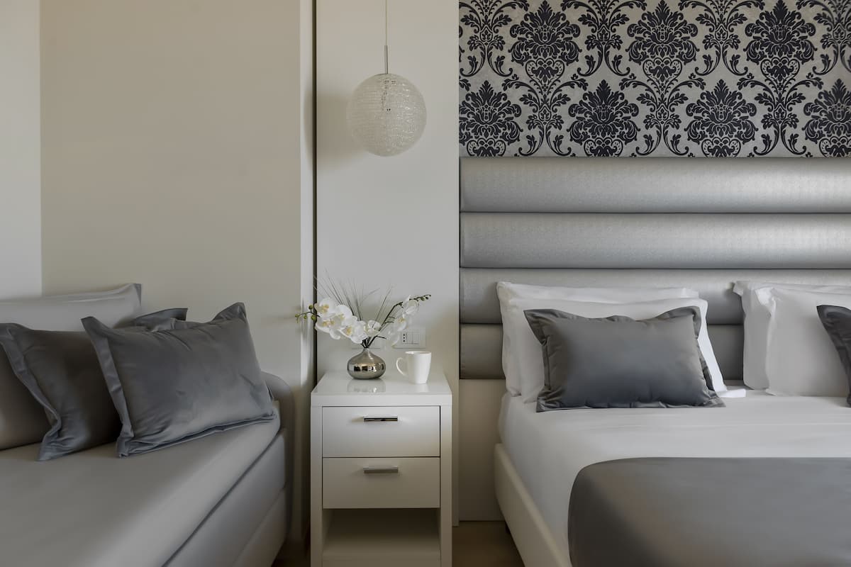 A modern bedroom with a grey sofa, a white nightstand with a vase of flowers, and a bed with grey bedding.