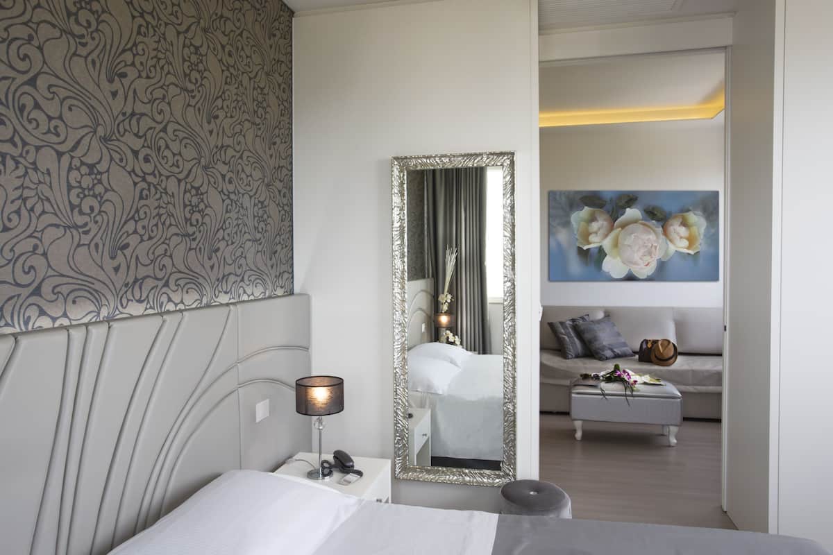 A modern hotel room with a large bed, a bedside table with a lamp, a mirror, and a painting of flowers on the wall.