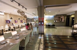 Un moderno vestíbulo de hotel con un separador de vidrio que divide el área de comedor de la recepción. Se observan mesas y sillas dispuestas con orden, un mostrador de recepción y una escalera que conduce a un nivel superior.