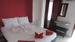 Una cama bien hecha con toallas blancas y rojas dispuestas en forma de lazo, una mesita de noche pequeña y un espejo en la pared.