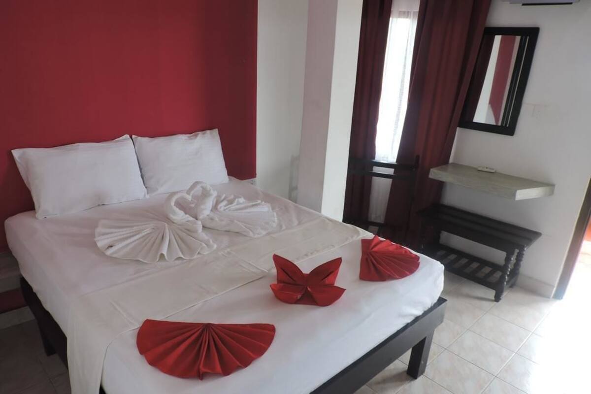 Una cama bien hecha con toallas blancas y rojas dispuestas en forma de lazo, una mesita de noche pequeña y un espejo en la pared.