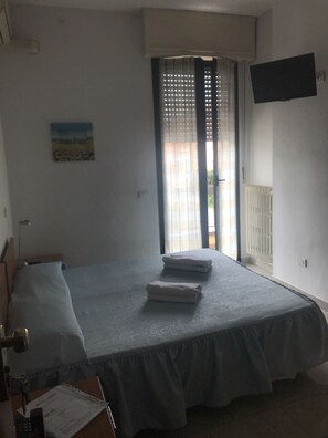 A single bed with a blue bedspread and two folded towels, a desk with a chair, a painting on the wall, and a TV mounted on the wall. - Hotel Cinzia (Savignano sul Panaro)
