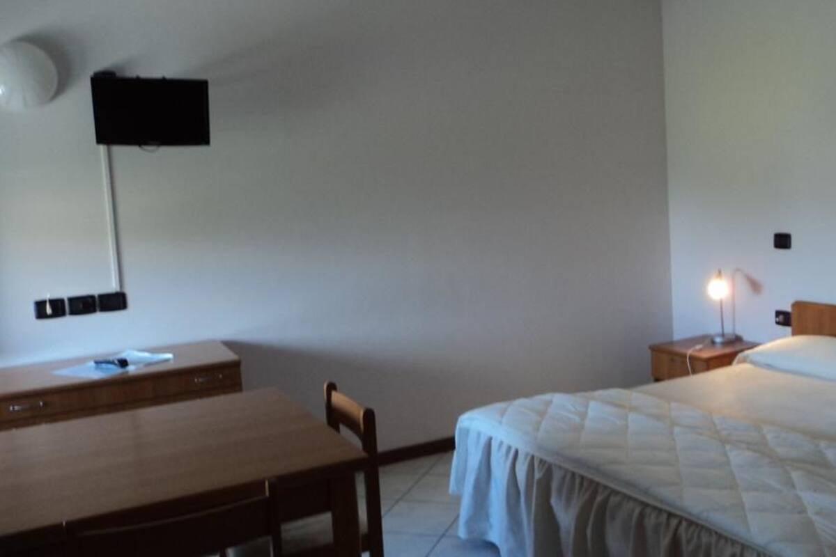 Een slaapkamer met een bed, nachtkastje, lamp, bureau, stoel en een aan de muur bevestigde televisie.