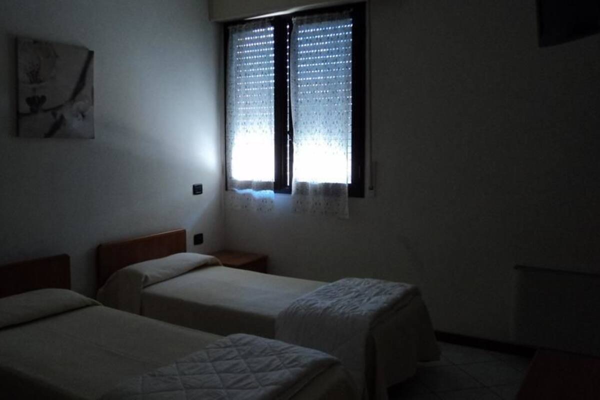 Een kamer met weinig licht, twee eenpersoonsbedden, een klein nachtkastje en een raam met dunne gordijnen.