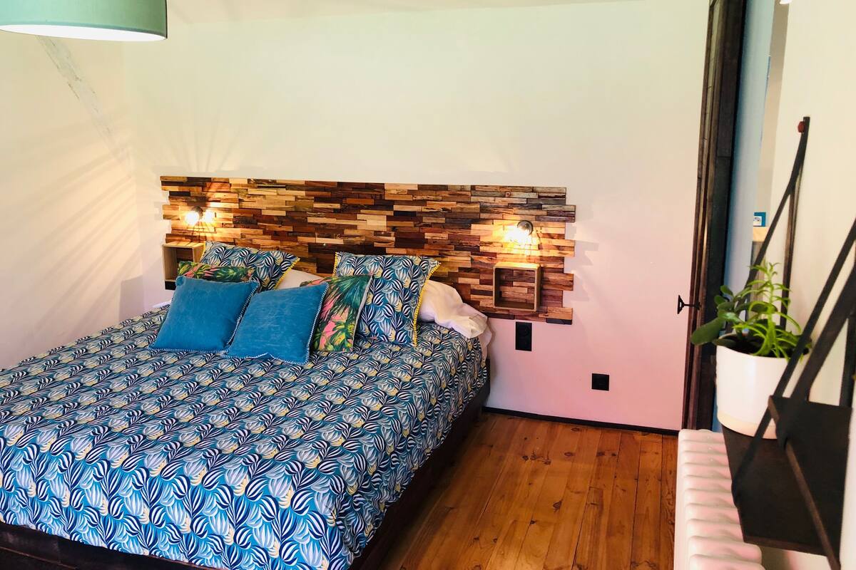 A bedroom with a bed, a wooden headboard, a hanging light fixture, and a potted plant.