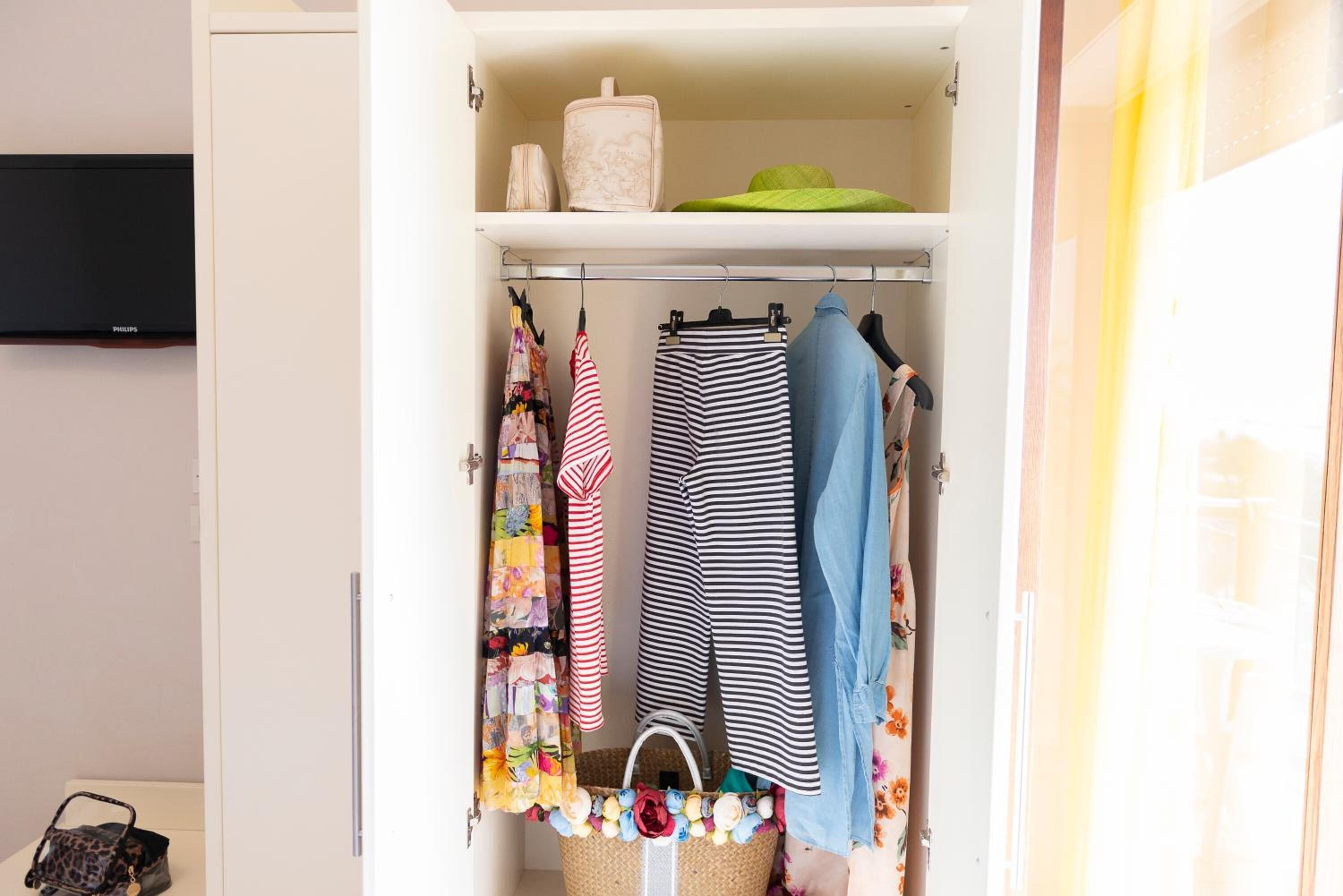 A closet with hanging clothes, a basket with flowers, and a television on the wall.