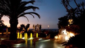 Une vue nocturne d’un complexe hôtelier avec une installation aquatique, des palmiers et une vue lointaine sur la mer.