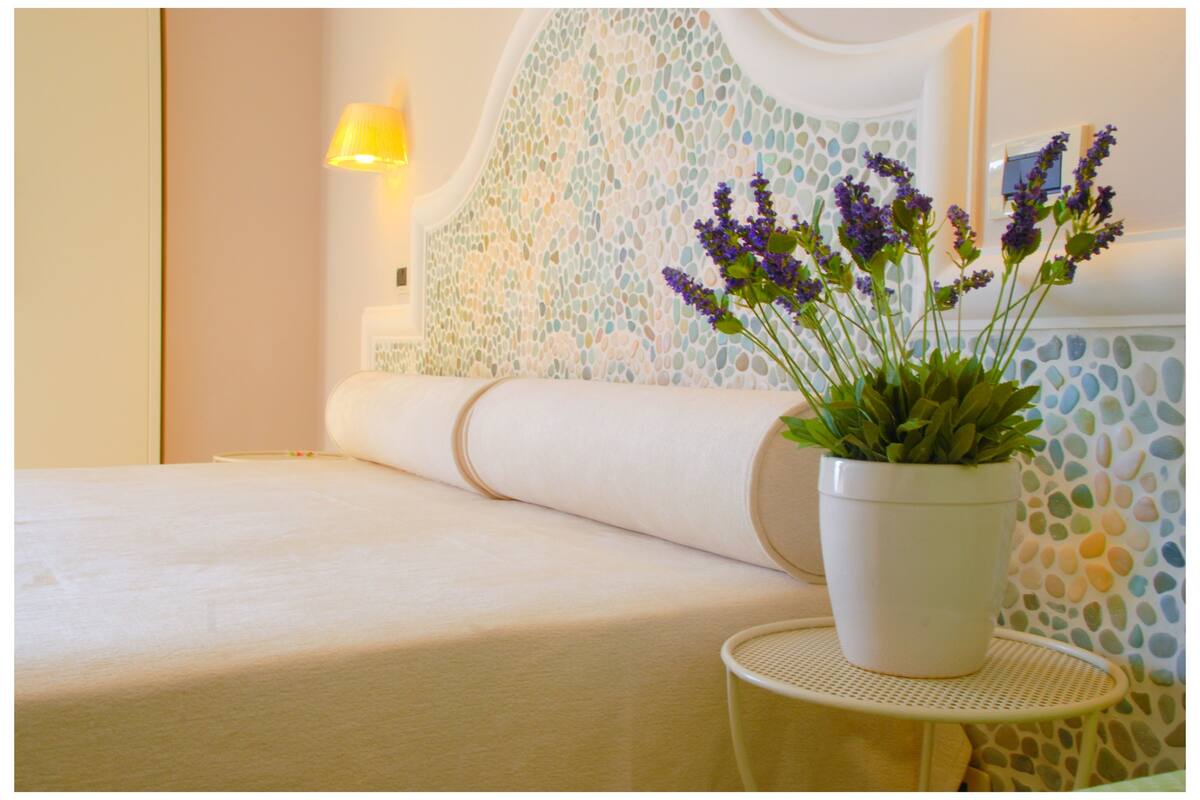 Un lit bien fait, recouvert d’une couette blanche, une petite table ronde sur laquelle est posé un pot blanc rempli de fleurs de lavande, et un mur orné d’un motif en mosaïque.