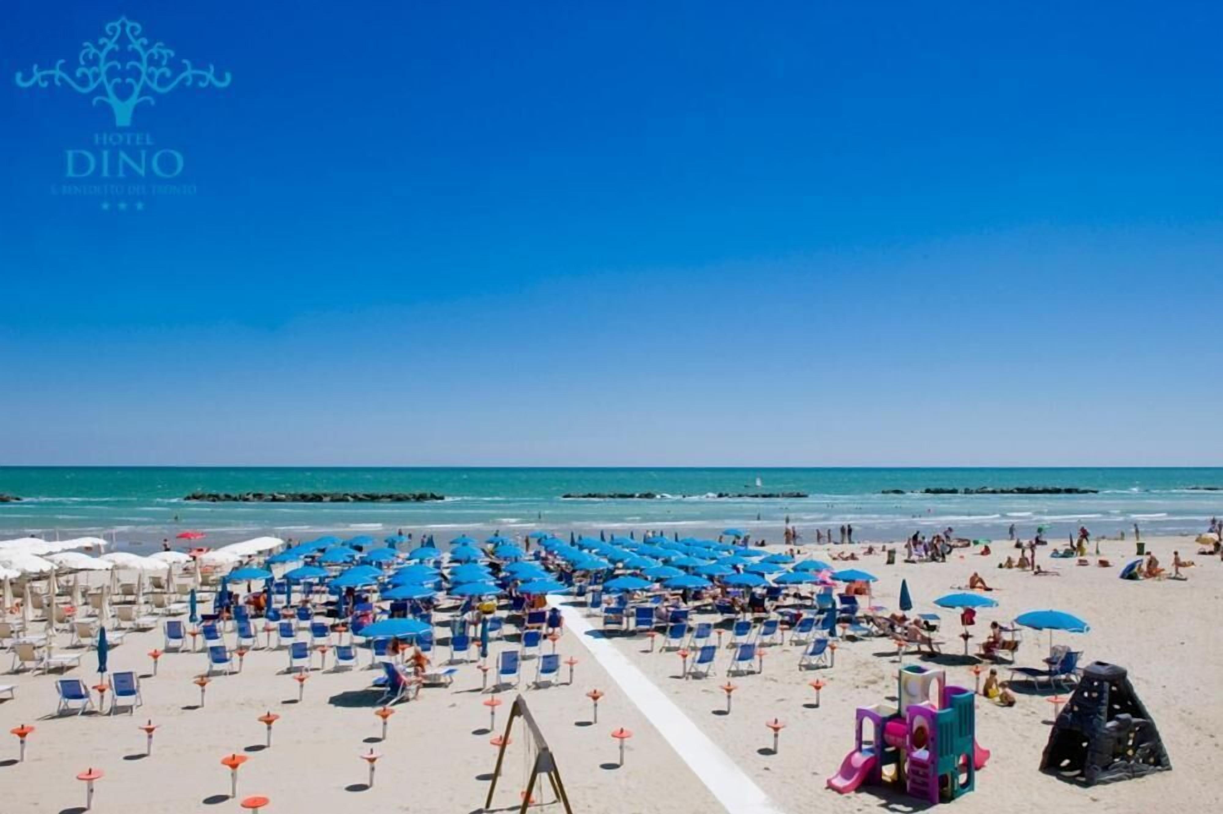 Una playa con hileras de sombrillas y sillas de playa azules y blancas, cielo despejado y el océano al fondo.