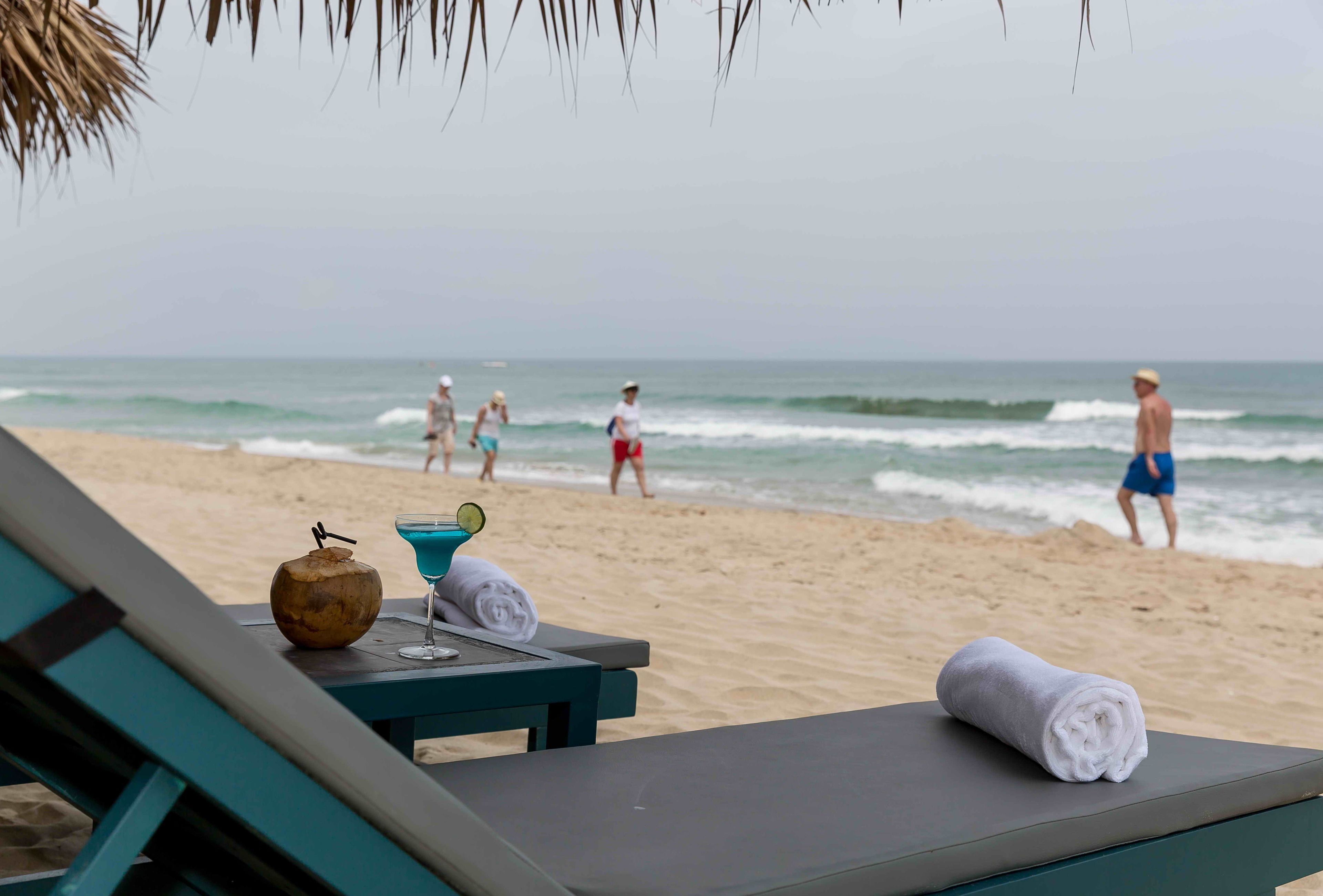 Blick auf einen Strand mit Liegestuhl, Handtuch, Getränk und Kokosnuss auf einem Tisch.