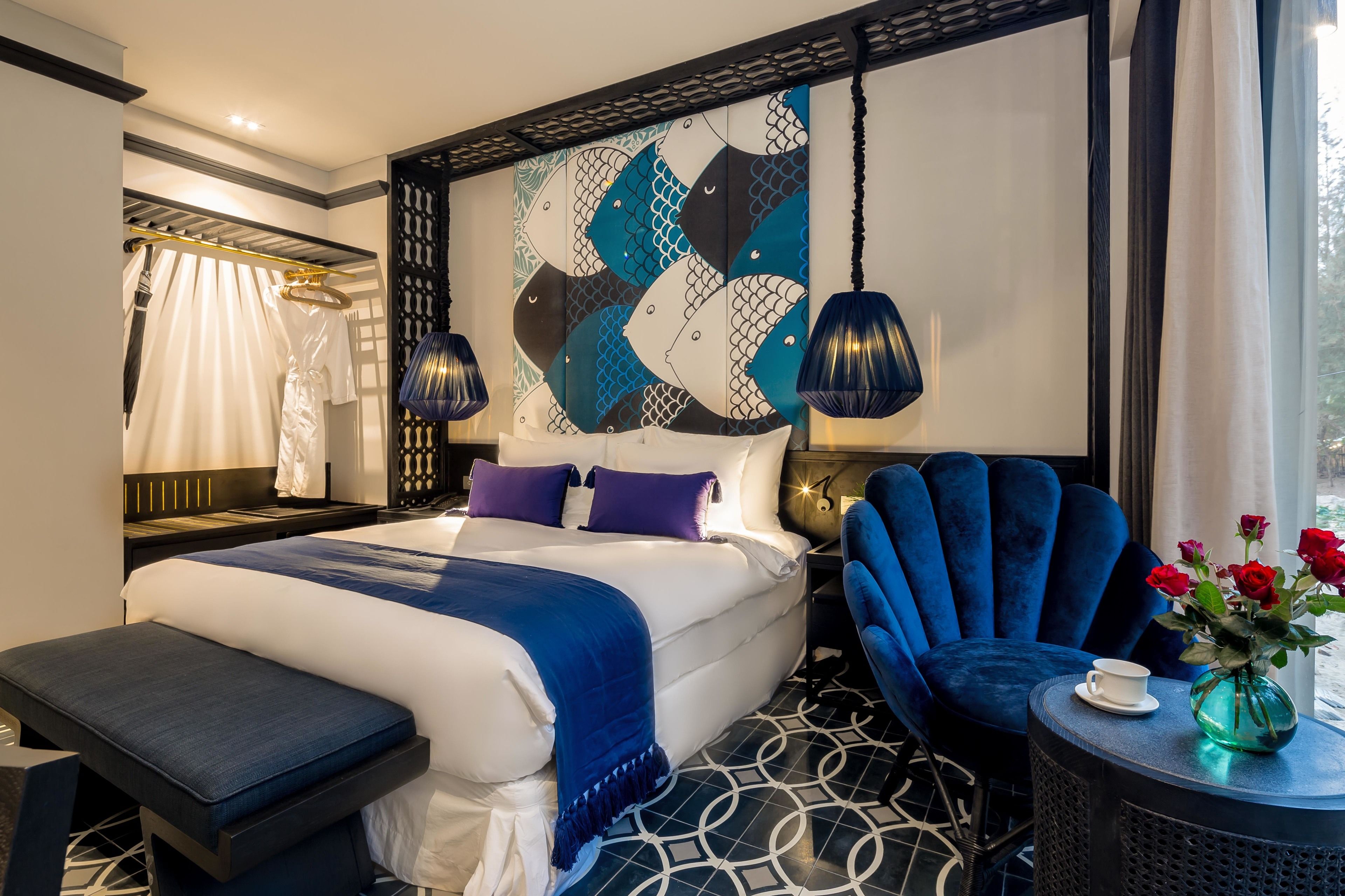 Une chambre d’hôtel moderne avec un grand lit, un fauteuil bleu, une table ronde ornée d’un vase de fleurs et un panneau décoratif mural.