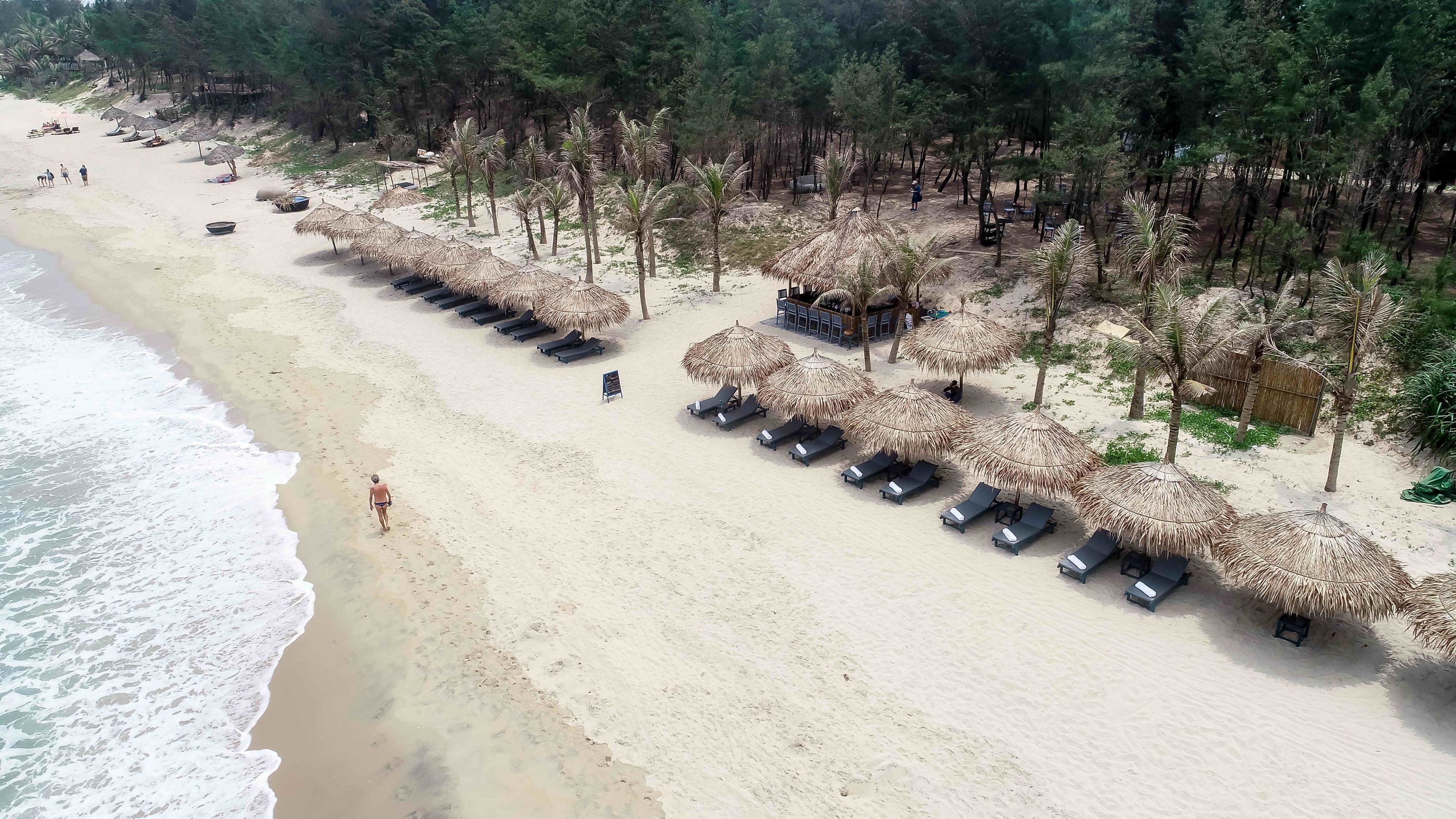 Una playa con sombrillas de paja, sillas de playa y algunas personas.