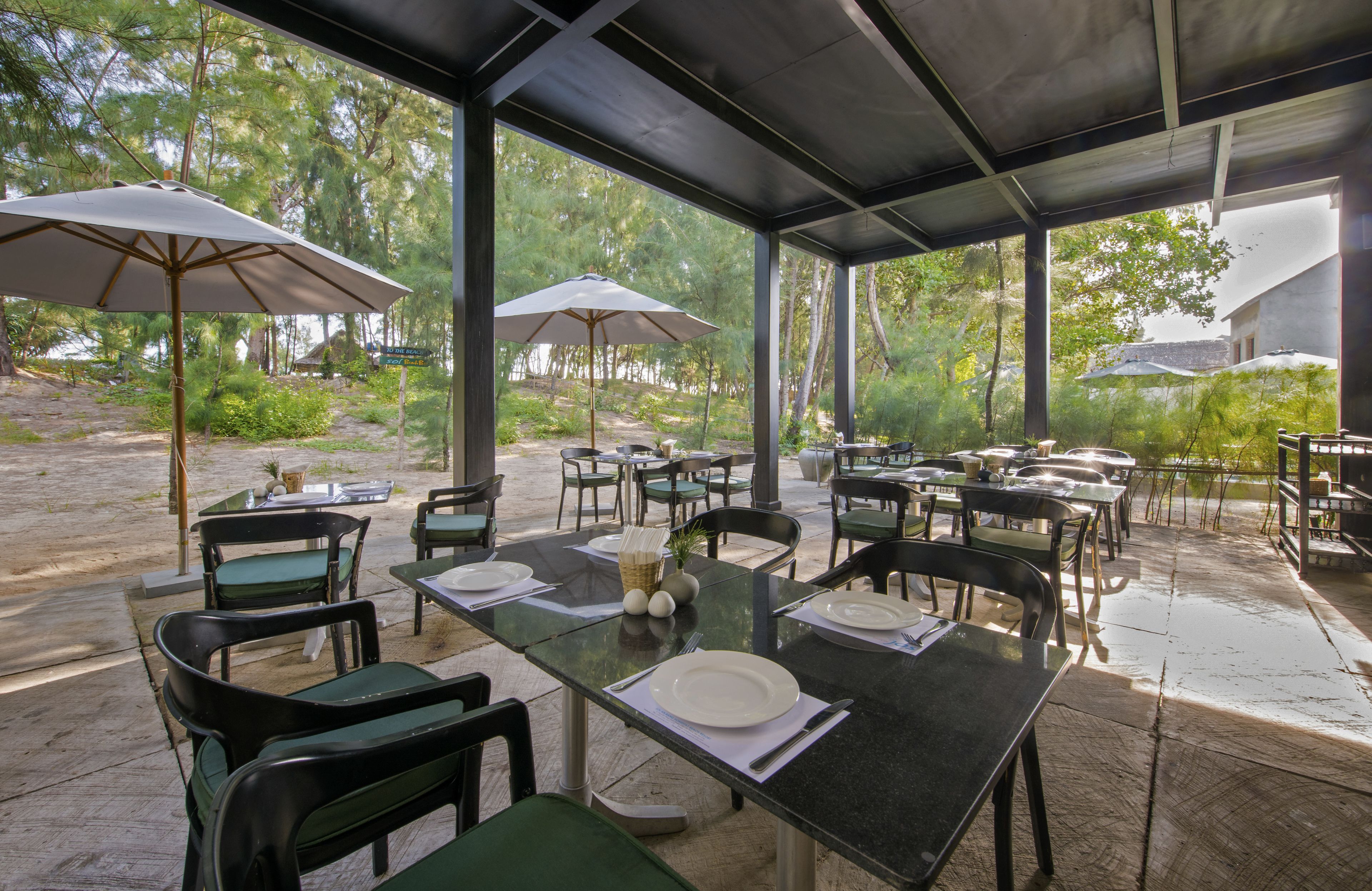 Un área de comedor al aire libre con mesas y sillas, sombrillas y vistas a árboles y un edificio al fondo.