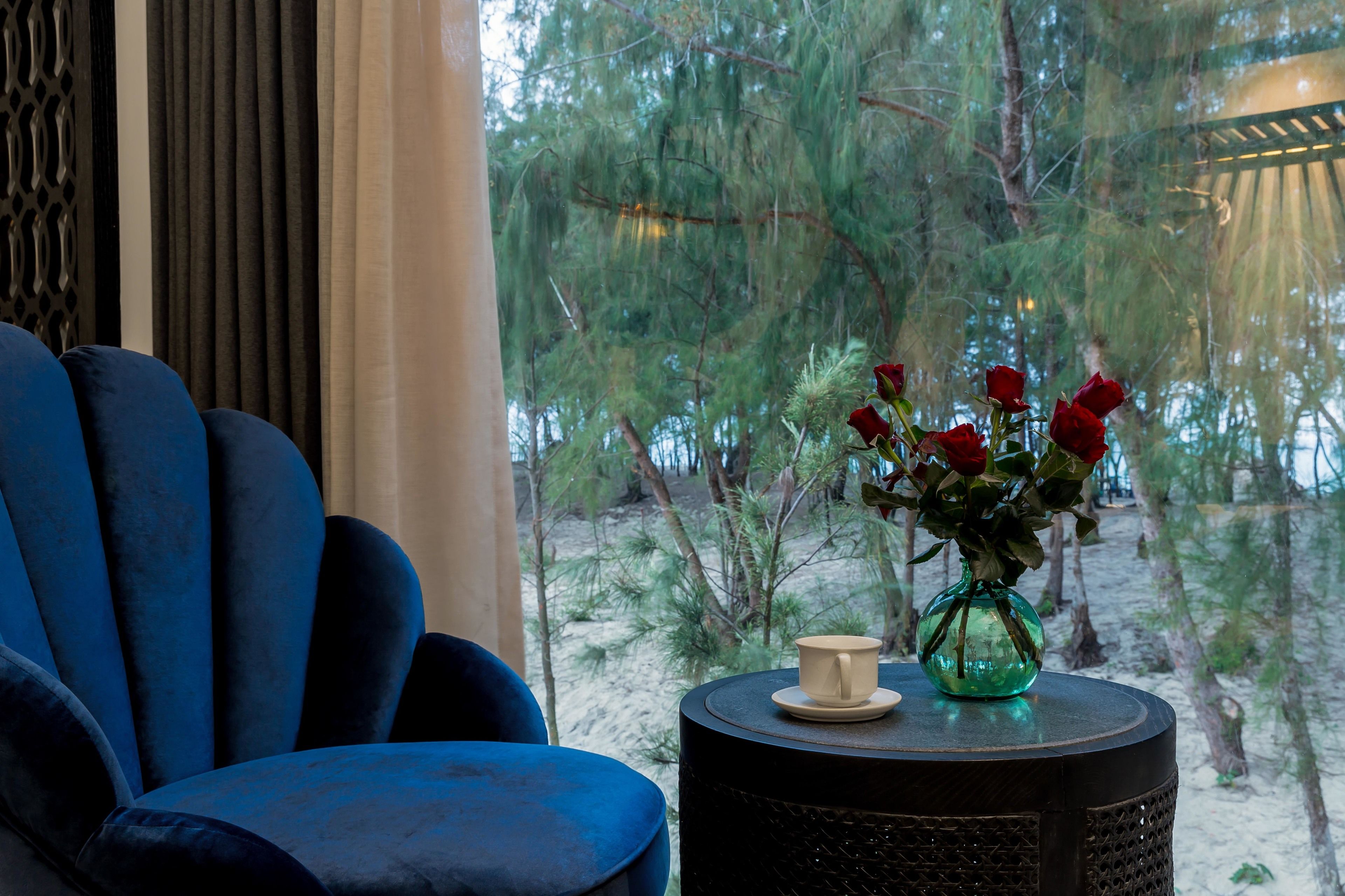 A blue armchair, a small round table with a vase of red roses and a cup, and a view of trees and sand through a window.
