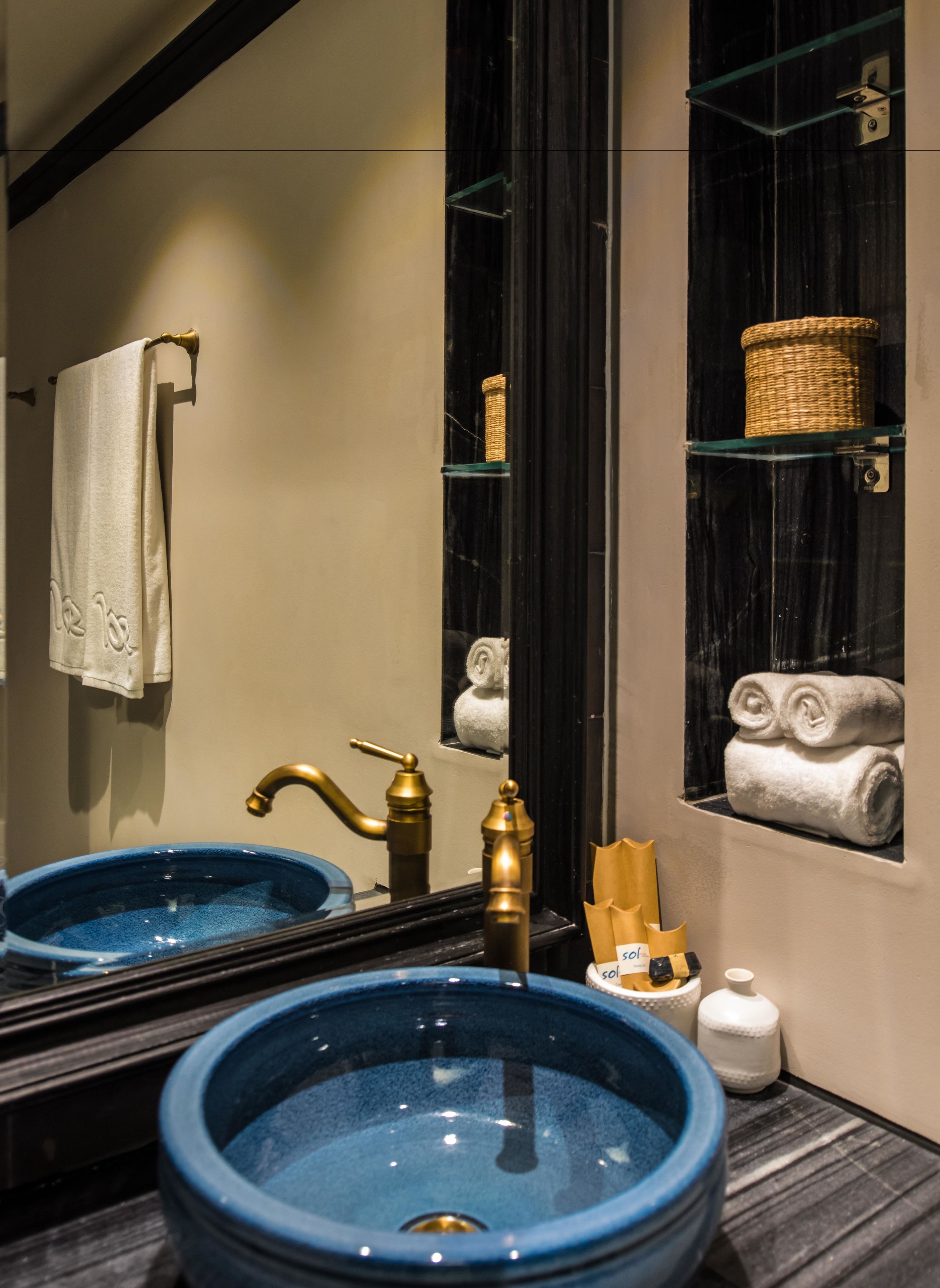 Un baño con dos lavamanos grandes de cerámica azul, un grifo dorado, un espejo y un estante con toallas y una cesta.