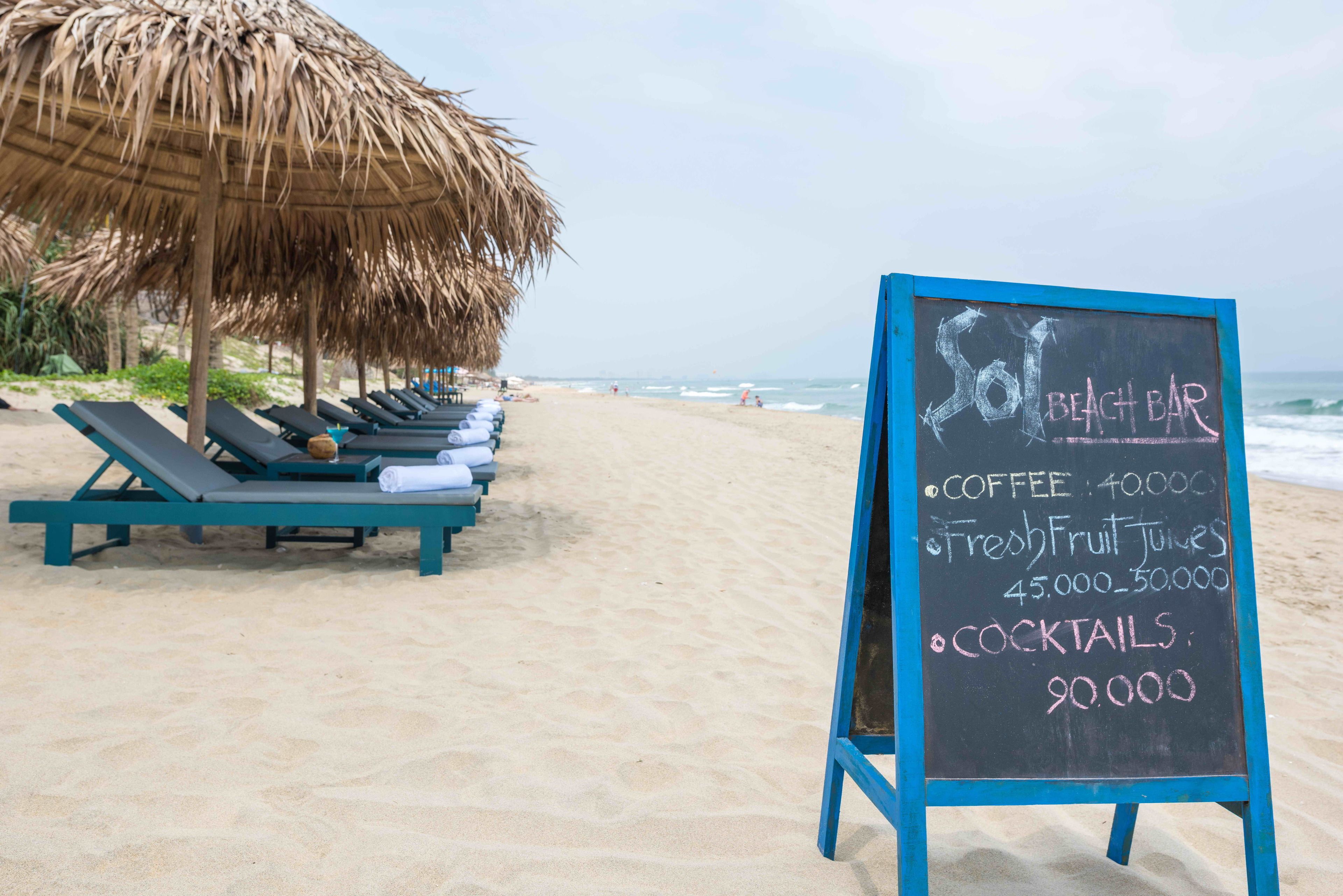 A beach bar with a menu board listing prices for coffee, fresh fruit juices, and cocktails.