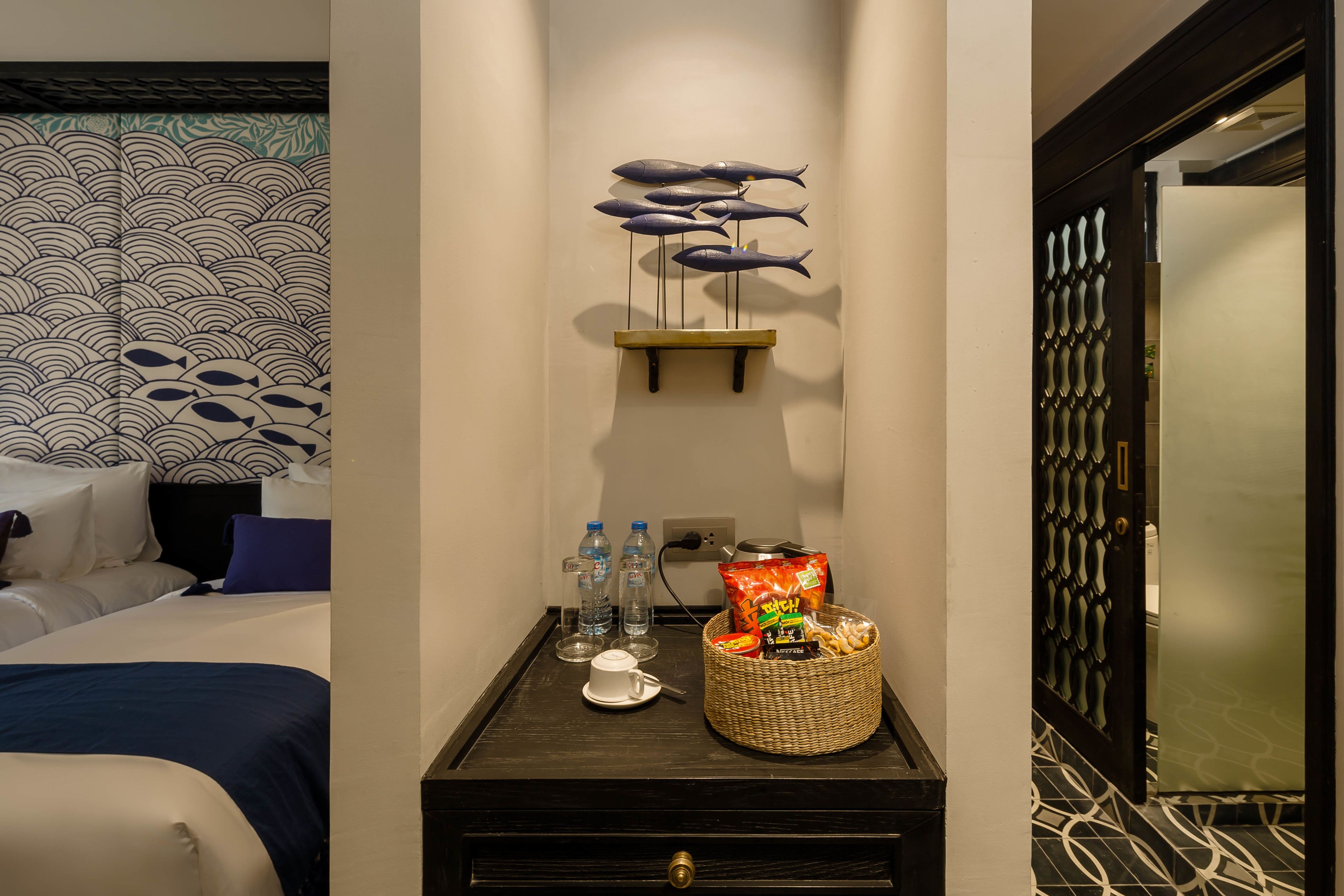 Habitación de hotel con una cama, una mesita con una cesta, una bandeja con aperitivos y una decoración de peces en la pared.