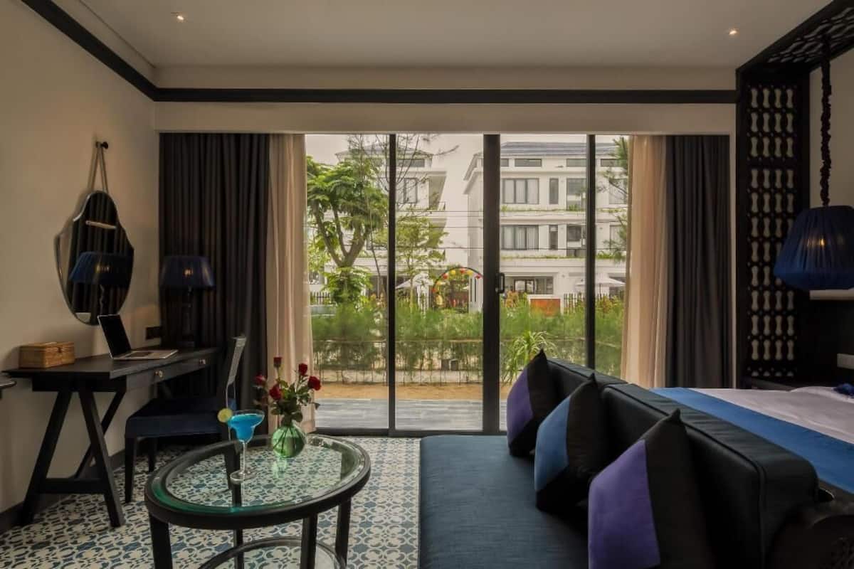 Habitación de hotel con cama, escritorio, silla, una mesita con un jarrón y vistas a través de un ventanal.