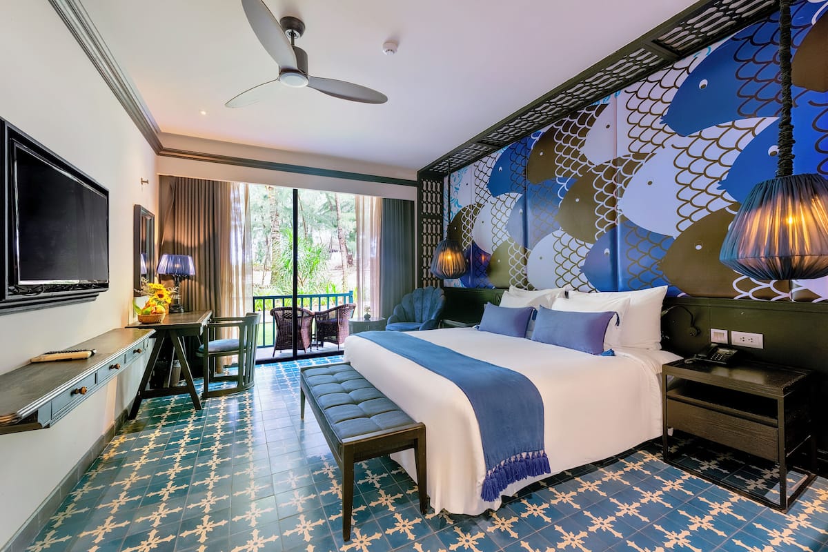 Habitación de hotel con una cama grande, un escritorio, un televisor y un balcón con vistas a la vegetación.