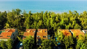 A coastal property with multiple buildings featuring red-tiled roofs, surrounded by dense green trees and a view of the sea.