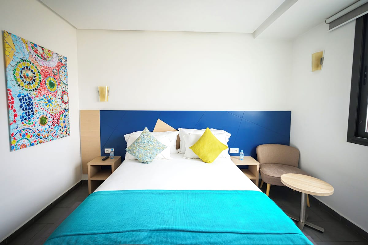 A hotel room with a large bed, a blue headboard, a colorful abstract painting, a small table, and a chair.