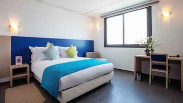 A modern hotel room with a large bed, a desk, and a window providing natural light.