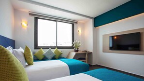 A hotel room with two beds, a TV mounted on the wall, a window with a view, and a small desk with a plant.