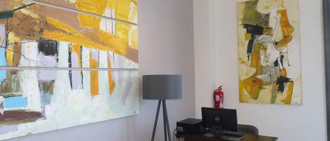 Une pièce dont les murs sont ornés de peintures abstraites, avec un bureau, un ordinateur, une chaise et un lampadaire.