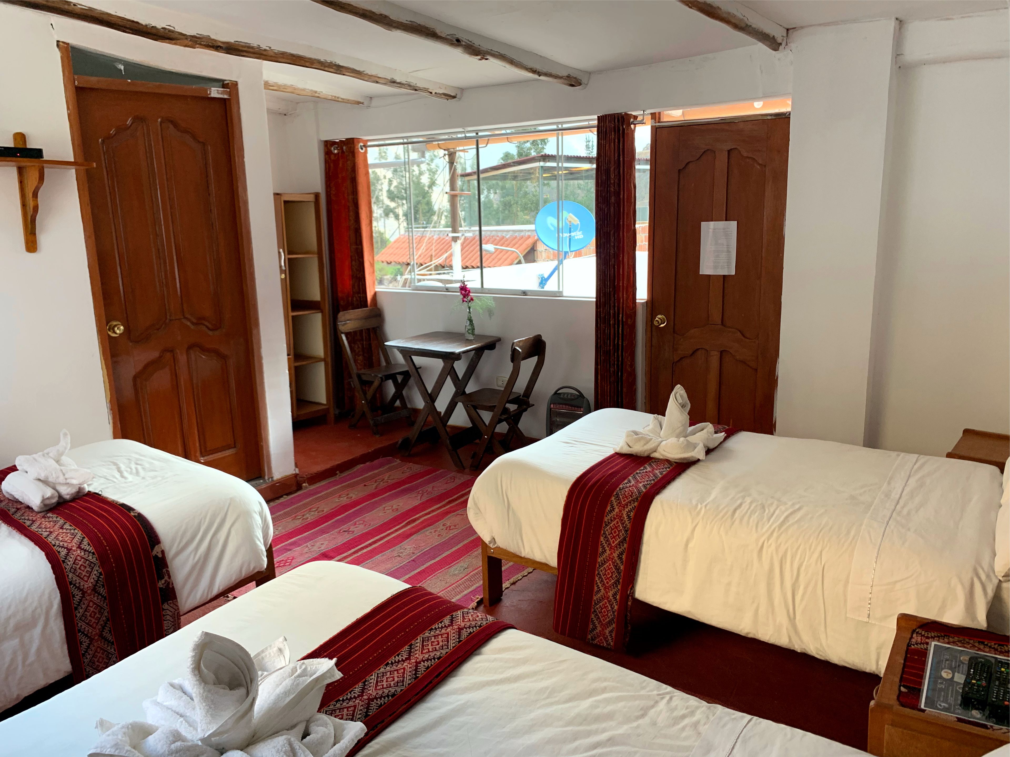 Habitación con dos camas, una mesita pequeña y una silla. Tiene una ventana con cortinas, una puerta y una estantería con objetos.