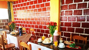 A breakfast setting with a brick wall, wooden chairs, a table with breakfast items, and a vase of flowers. - Inka Khawarina Tambo Lodge (Ollantaytambo)