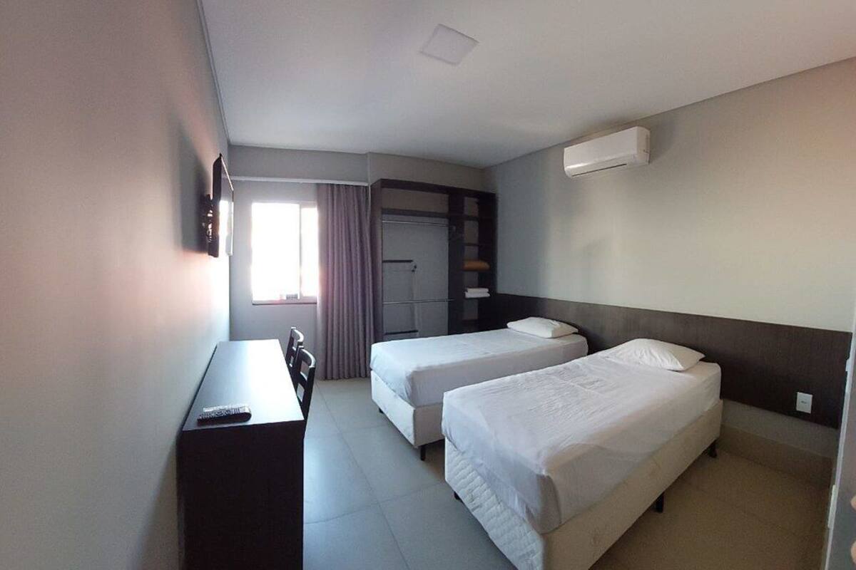 A hotel room with two single beds, a desk, a chair, a wardrobe, and a window with curtains.