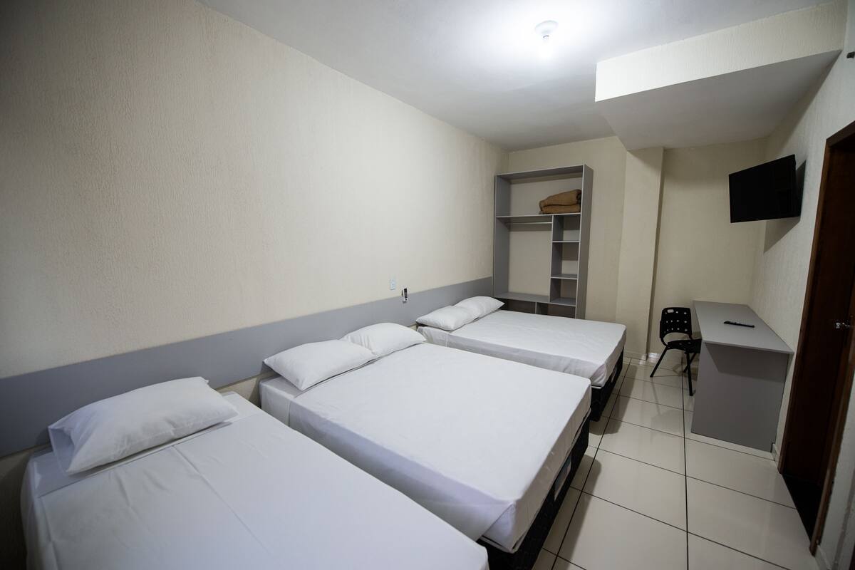 A hotel room with three single beds, a desk, a chair, a TV mounted on the wall, and a wardrobe.