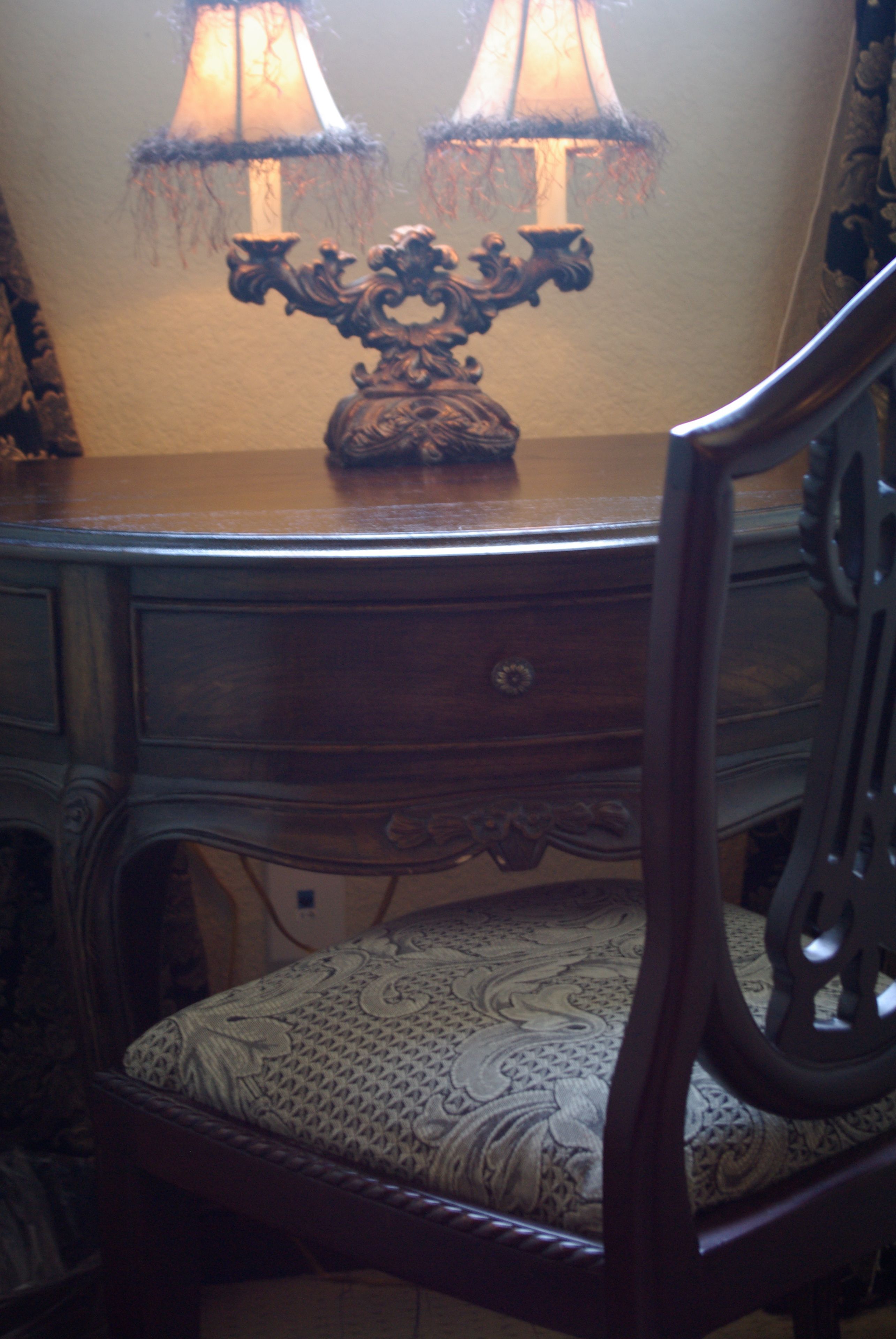 Un bureau en bois avec deux lampes, une chaise dotée d’un coussin à motifs et un miroir qui reflète les lampes.