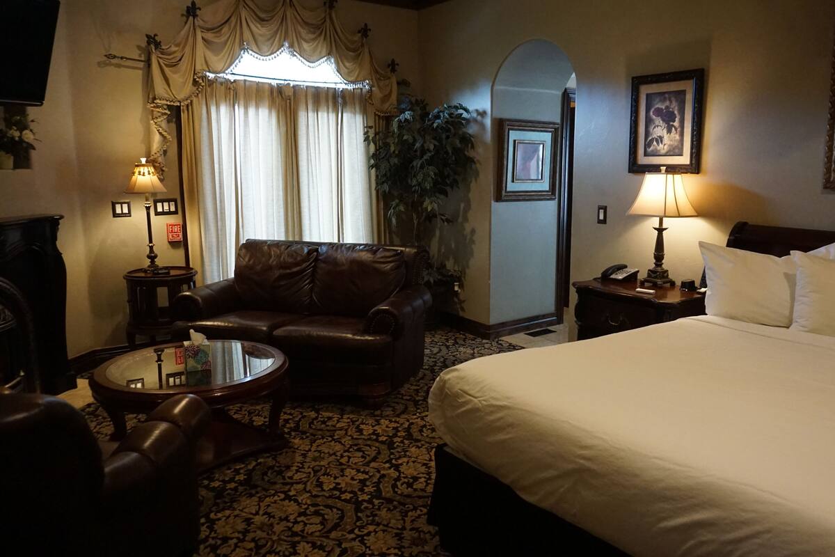 A hotel room with a large bed, a leather armchair, a small round table, a fireplace, a large window with curtains, a painting, and a lamp.