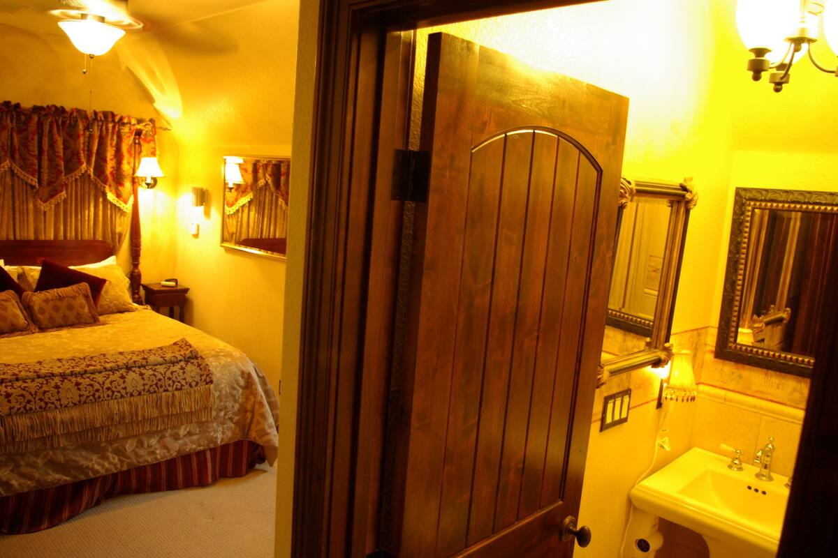A hotel room with a bed, a wooden door, a bathroom with a sink and mirror, and a chandelier.