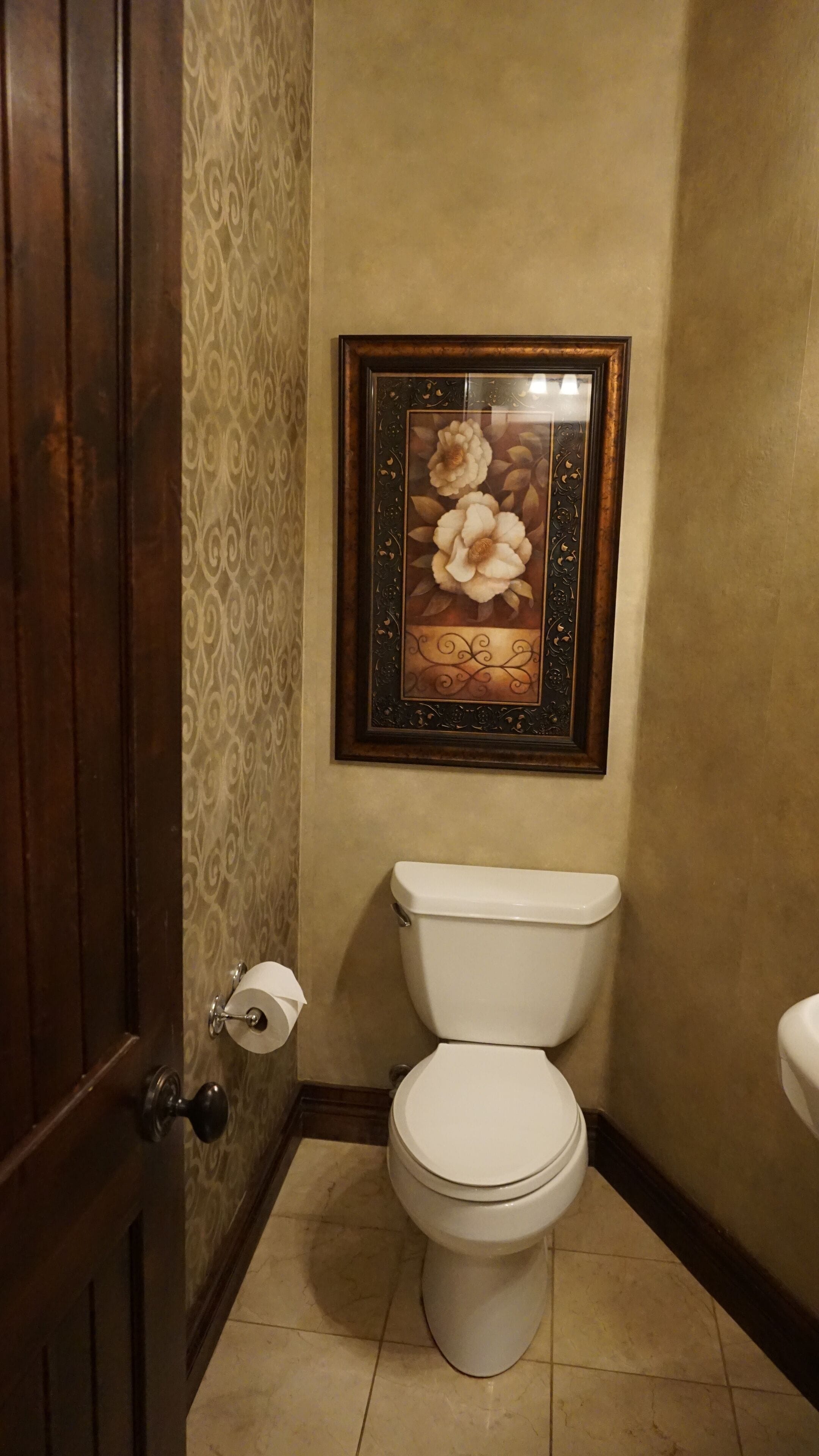 A bathroom with a toilet, a framed floral artwork on the wall, and a patterned wallpaper.