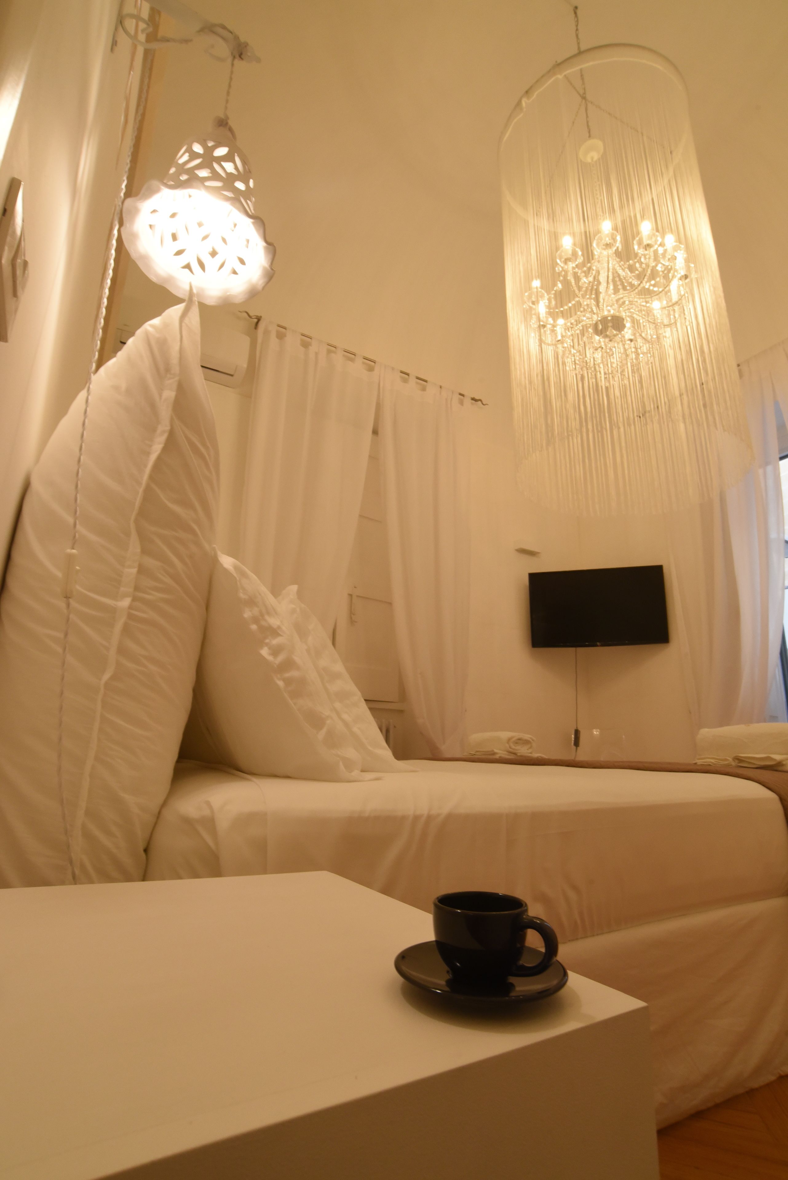 A hotel room with a bed, a bedside table with a cup, a TV mounted on the wall, and a chandelier.